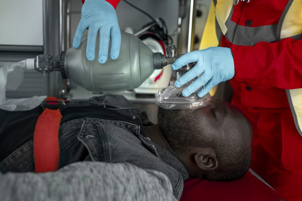 Image of a man lying down with his eyes closed and given an emergency oxygen mask by a First Responder
