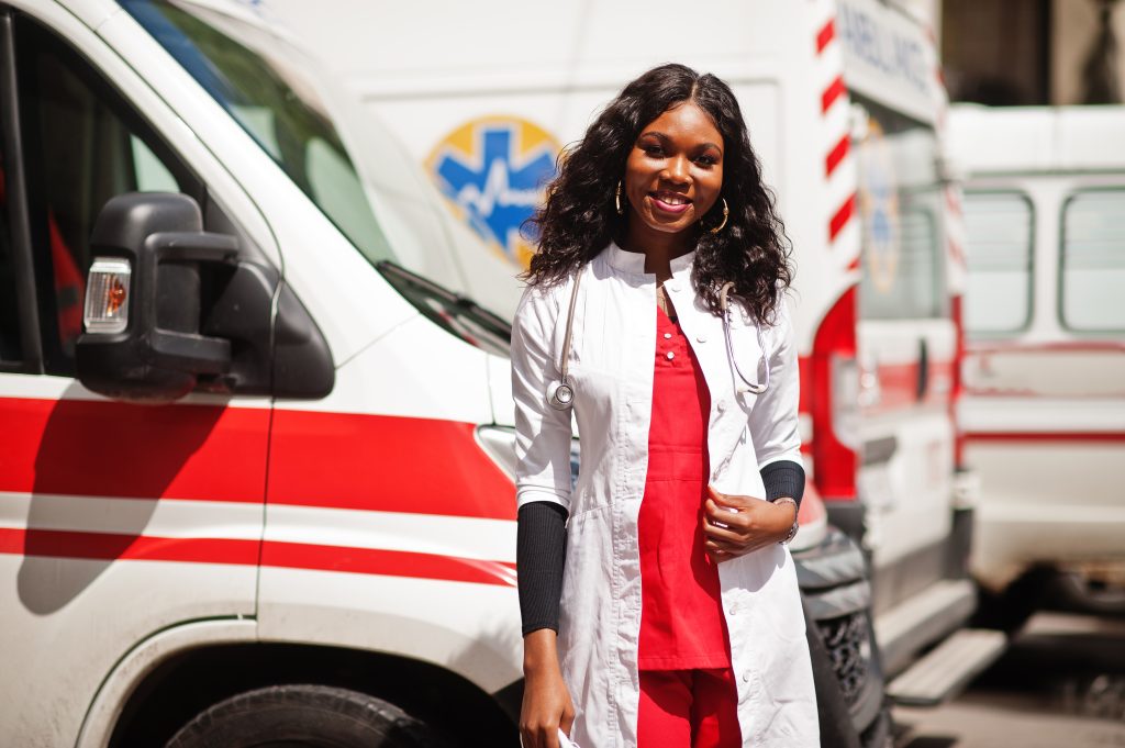 Image of a smiling black girl in front of an ambulance wearing red scrubs and a white coat